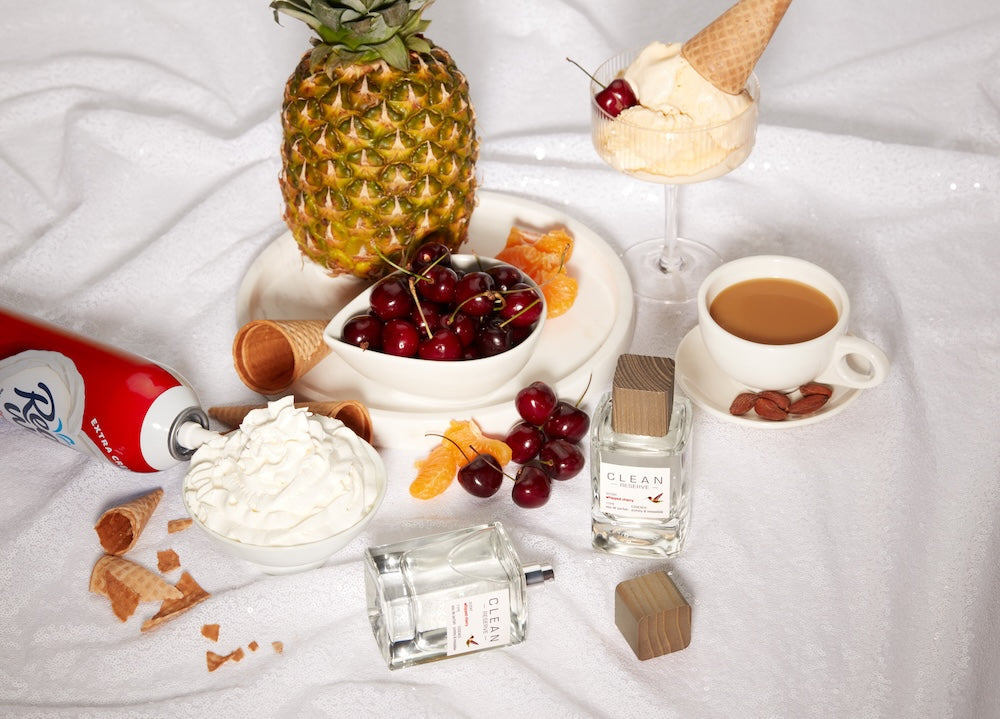 Flat lay arrangement featuring cherries, pineapple, ice cream, whipped cream, coffee, waffle cones, citrus slices, and CLEAN perfume bottles on a soft white fabric background, styled with a cozy dessert-inspired aesthetic.