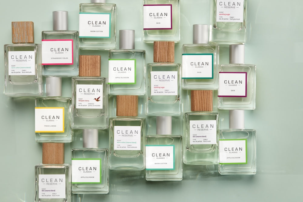 Collection of CLEAN perfume bottles arranged on a soft green background, featuring minimalist packaging with colorful labels and wooden or metallic caps in a modern flat lay display.