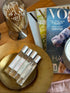 Three small clear glass perfume bottles with silver caps and white labels are arranged on a round gold tray beside a glass jar of matches and a magazine on a wooden table.